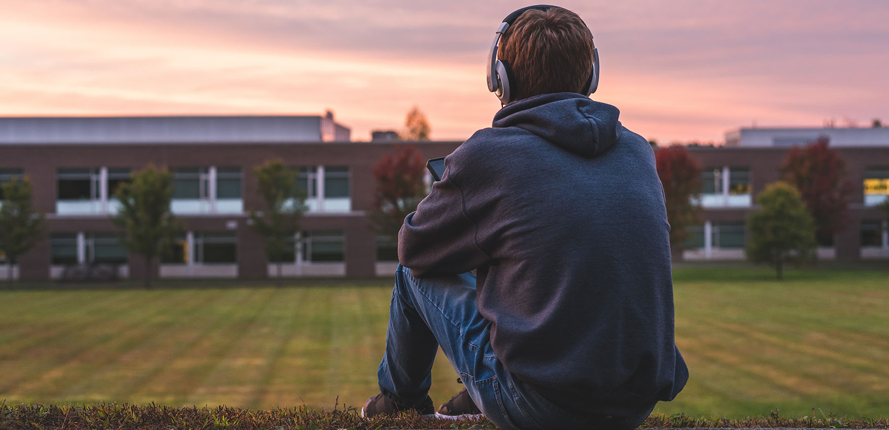 a teenaged boy wearing headphones sits on a path looking at a school building