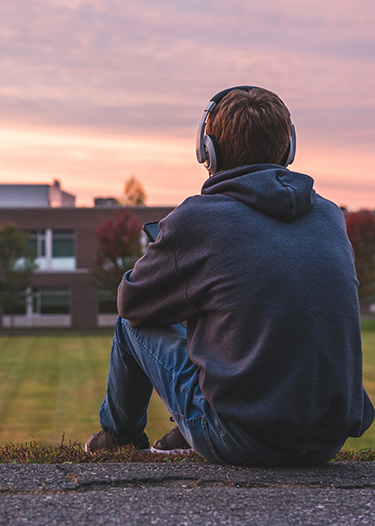 a teenaged boy wearing headphones sits on a path looking at a school building