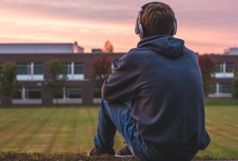 a teenaged boy wearing headphones sits on a path looking at a school building